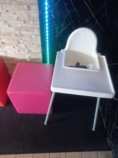 An indoor scene features a white plastic baby high chair positioned to the right of a pink upholstered cube ottoman. The high chair, with light grey metal legs and a white tray, contains visible black safety straps. Both objects rest on a dark, textured mat that partially covers light-colored wooden floorboards. The background consists of a wall with light, horizontal stacked stone veneer on the left, and a dark, reflective wall with white marble-like veining on the right. A vertical strip of illuminated blue-green LED lights is affixed to the right wall between the two distinct wall sections. No individuals are present. The image does not contain details to confirm its location in the city of Sallaumines, France.