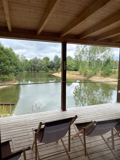 The image depicts an outdoor seating area overlooking a lake surrounded by trees. In the foreground, there is a wooden deck with several canvas lounge chairs. The chairs are arranged facing the water. A thick rope runs horizontally across the deck, separating it from the lake. The lake itself is calm and reflects the cloudy sky and the surrounding greenery. Beyond the lake, there is a sandy shore and a dense forest. The architecture of the seating area is rustic, with exposed wooden beams forming the ceiling. The overall scene suggests a tranquil, natural setting, likely a recreational area by a body of water. This location appears to be within the city of Dobrzyń, Poland, given the context provided.