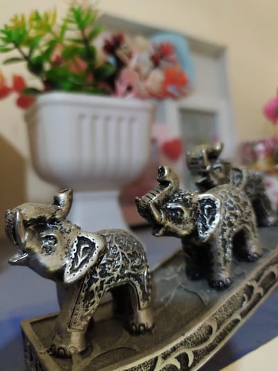 A close-up, shallow depth of field shot shows a row of four small, metallic elephant figurines. The elephants are facing to the left, with intricate patterns carved into their bodies. The foreground features the leftmost elephant prominently, appearing to stand on a decorative base. Behind it, three other elephants are visible in decreasing size and focus. In the background, out of focus, is a white planter containing artificial flowers and foliage in shades of green, pink, and orange. The overall setting appears to be an indoor environment, likely a shelf or tabletop. The location is not specified as Bilajari, Azerbaijan.
