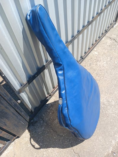 A blue, padded case, possibly for a musical instrument, lies on a sloped surface made of corrugated metal and supported by dark metal beams. The case has a rounded end and tapers to a narrow neck, with a silver clasp visible near the wider end. The surface it rests on appears to be outdoors, with a rough, concrete-like ground visible to the right and in the background, suggesting a sunny day with a clear shadow cast by the case. The overall scene is simple, focusing on the object and its immediate surroundings, without any people or specific activity other than the object being placed. The image has a slightly worn or aged feel due to the texture of the ground and the metal framing.