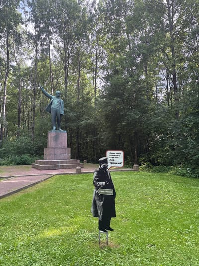 In Sestroretsk, Russia, a statue of Vladimir Lenin stands on a stone pedestal in a grassy clearing surrounded by tall trees. In the foreground, a cardboard cutout of a man in historical attire, identified as Lev Trotsky by a nearby sign, stands on the grass. The sign, shaped like a speech bubble, asks in Russian, "Lenin hid here, where was Lev Trotsky?". A smaller sign below the Trotsky cutout reads "answer in the museum". The environment is a park or wooded area with dappled sunlight on the grass.