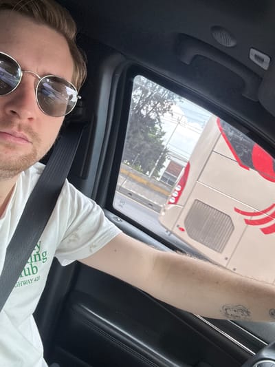 A male with blond hair and a light beard, wearing sunglasses, a white t-shirt with green text that reads "HIGHWAY 420", and a black seatbelt, is driving a vehicle. He is looking at the camera. Through the car window, a beige and red bus is visible on a road. The surrounding environment appears to be an urban or suburban area with trees and buildings. The scene is captured from inside the car, suggesting a personal photo taken while traveling. The image is dated and located in Cuautitlán Izcalli, Mexico.