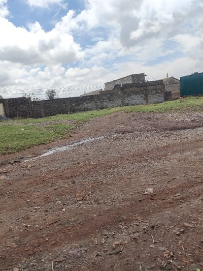 The scene depicts an unpaved outdoor area in Juja ward, Kenya, under a sky with extensive white and grey cloud cover. The foreground consists of a reddish-brown dirt path, uneven and scattered with small aggregate and debris, including a discarded white object and twigs. A distinct linear puddle of water is present along the left side of the dirt path. Adjacent to the path on the left is a strip of green grass. A dark grey concrete block perimeter wall, exhibiting varied construction and height, extends horizontally across the midground. Behind the wall, several multi-story structures constructed from light-colored concrete blocks are visible, appearing unfinished. To the far right, a building features a dark green/teal corrugated metal wall section. Overhead utility lines are visible traversing the mid-to-background against the sky. No human figures or discernible activities are observed.