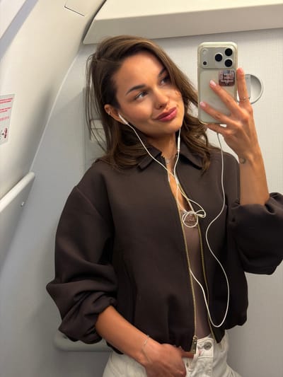 A woman is taking a selfie with her iPhone, likely inside an airplane. She is wearing white earbuds, a brown jacket with a gold zipper, a sheer top, and white pants. She has brown hair and is wearing rings on her fingers. The setting appears to be Емельяново (аэропорт), Russia, as evidenced by the typical airplane interior.