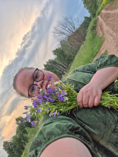 In a selfie taken from a low angle, a person with blonde hair and glasses is holding a bouquet of small purple and yellow wildflowers. The person is wearing a green dress with a floral pattern. They are outdoors, with a dirt road and grassy fields visible in the background. Trees and a cloudy sky with hints of sunset are also present. The overall scene suggests a rural or natural setting, possibly in or around Doinaya, Russia, given the context.
