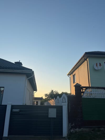 This is a rotated photograph of two houses and the sky. The sky is clear and blue with a warm glow on the horizon, suggesting it is either sunrise or sunset. A small bird is visible flying in the upper left portion of the sky.

The foreground is dominated by the roof and upper facade of a modern white house with a dark gray, textured roof. The house has large windows. Adjacent to it, and partially obscuring it, is a dark gray corrugated metal gate or wall.

In the upper portion of the image, a light green house with white trim is visible. It has horizontal siding and a satellite dish mounted on its exterior. A white picket fence is also visible. Between the two houses, a white fence with decorative cutouts can be seen, along with dark green vertical siding.

The scene depicts a residential area. The lighting indicates it is either early morning or late evening. There is no activity or people visible in the image. No text is discernible. The location is identified as Navahrudak, Belarus.