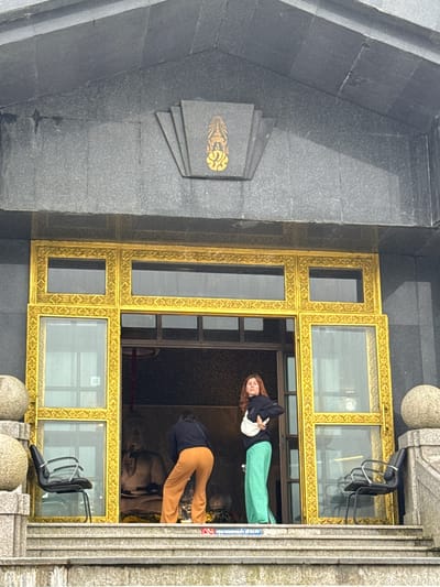 The image shows the entrance to a building with a large, ornate golden frame around the doorway. The building's facade is made of dark granite. Two women are present. One woman, wearing a dark top and orange pants, is inside the doorway, facing away from the camera and appears to be interacting with something within the building. The other woman, with red hair, wearing a dark jacket and green pants, stands in the doorway, facing the camera. To the left and right of the entrance, black chairs are placed. The steps leading up to the entrance are made of stone. A Thai royal emblem is visible above the doorway. The overall scene appears to be a religious or cultural site, possibly in Chang Khoeng, Thailand.