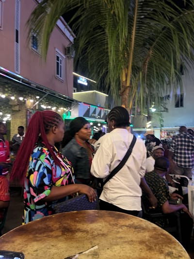The image captures a nighttime scene at a vibrant outdoor venue in Oba, Nigeria, likely a restaurant or lounge. The foreground is dominated by a large, textured wooden table, hinting at a communal gathering space. In the mid-ground, several individuals are engaged in conversation or observing their surroundings.

A woman with a distinctive long, braided red ponytail is prominently featured on the left, wearing a colorful, abstract-patterned top. She is looking to her right, engaged in conversation with another woman. This second woman, with dark hair, is wearing a denim jacket over a red top.

Further to the right, a person wearing a white button-down shirt and dark pants, with a black bag slung across their chest, is facing away from the camera, also appearing to be in conversation. Several other individuals are visible in the background, some seated and others standing, amidst the ambient lighting of the establishment.

The environment is characterized by lush palm fronds from a large tree that partially obscures the view, suggesting a tropical or sub-tropical setting. Overhead, strings of lights are visible, contributing to the festive atmosphere. The architecture of the building in the background, with its pink facade and windows, indicates an urban setting. Neon signage, partially visible, adds to the nighttime allure of the location. The overall impression is one of a lively social gathering.