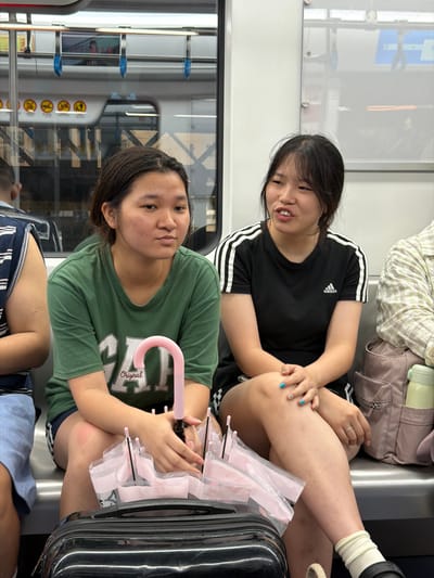 The image shows two young Asian women sitting on a train. The woman in the foreground is wearing a green t-shirt and dark shorts. She has dark hair and is looking towards the camera with a neutral expression. She is holding a pink umbrella with a translucent pink cover. A suitcase is next to her. The woman in the background is wearing a black Adidas t-shirt and dark shorts. She has dark hair and is looking away from the camera, smiling slightly. She has a ring on her left hand. A backpack is behind her. The setting appears to be a public transportation vehicle, possibly a train, with windows showing a blurred urban or rural landscape outside. The lighting suggests it could be daytime. The overall mood is casual and relaxed.