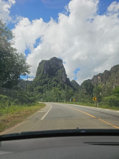 This media file depicts a scenic view from a car traveling along a curved road in Ban Nai Sa, Thailand. The scene is dominated by lush green mountains on the left, covered in dense vegetation and dramatic rock formations. The road itself is asphalt, with a yellow line marking the center and white lines delineating the lanes.

The sky is a vibrant blue, with large, puffy white clouds scattered across it, suggesting it is a sunny day, possibly mid-morning or afternoon. The sunlight casts shadows, indicating a bright day. The presence of power lines with poles suggests a developed area, even within the natural landscape.

On the roadside, there are signs with yellow chevron arrows, indicating curves in the road. The vegetation along the roadside is thick and green, consisting of grass and trees. The overall impression is one of a beautiful, natural environment with well-maintained infrastructure. There are no people or specific objects that draw significant attention, the focus being on the expansive landscape and the road that winds through it.