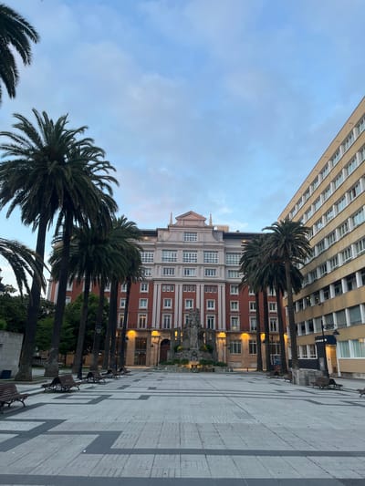 The image shows a street scene in La Coruña, Spain. Tall buildings flank a pedestrian square paved with gray and white stones. A row of palm trees lines one side of the square, interspersed with benches and lampposts. The buildings exhibit a mix of architectural styles, with one appearing modern and another with a red-brick facade and white window frames. The sky is overcast, suggesting either dawn or dusk. No people are visible in the immediate scene.