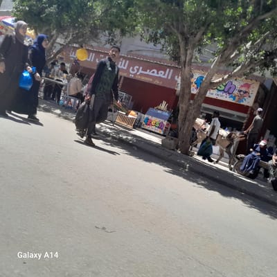 The image shows a street scene in Deir el-Balah, Palestinian Territories. In the foreground, a person in dark clothing walks on the street, carrying a blue bag. Further down the street, a man in a green and black athletic shirt walks with a backpack. To the right, a horse-drawn cart is present, with a man leading the horse and another person seated on the cart. Several other individuals are visible in the background, some near shops. Trees line the street, casting shadows on the ground. Signs for businesses are visible on the buildings. The overall scene depicts a typical day on a street in the city, with people going about their activities.