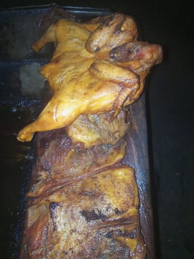 A whole roasted chicken rests atop a wooden board, accompanied by sizable, cooked meat pieces underneath. The chicken is golden-brown, with visible legs and wings. The meat below appears well-done and slightly charred. The scene is darkly lit, possibly indoors, emphasizing the food's colors and textures. A dark tray partially visible at the left suggests a preparation area.