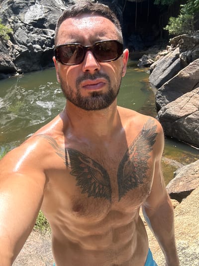 A man with sunglasses and a chest tattoo stands in front of a waterfall. The man is shirtless and appears to be sweating. The background is a rocky landscape with lush greenery and a body of water. The lighting suggests it is daytime, with sunlight casting shadows on the man and rocks.