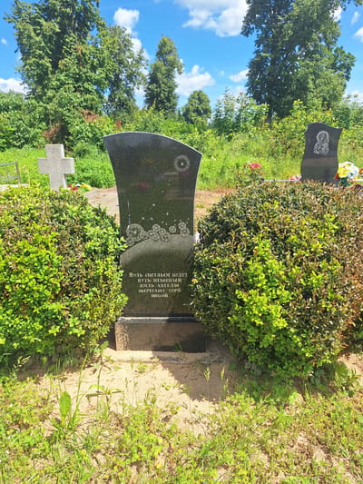 The image depicts a cemetery in the village of Belyevichi, within the Belyevichi rural council of Belarus. The scene is dominated by a black granite headstone, likely a gravestone, standing upright. The headstone has a curved top and is engraved with text and a circular design featuring stylized dandelion seeds. To the left of the headstone, a portion of a gray stone cross is visible. Two large, manicured green bushes flank the headstone, one on each side, partially obscuring the base of the gravestone and the surrounding ground. The ground in front of the headstone is sandy with sparse, green vegetation. In the background, a line of trees forms a dense green backdrop under a bright blue sky with scattered white clouds. Another, smaller headstone with an etched religious image is visible to the right, behind the right-hand bush, along with some colorful flowers, suggesting recent placement. The overall impression is that of a well-maintained, serene burial site on a sunny day.