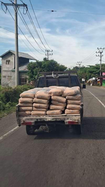 A pickup truck is captured from behind, driving on a road, presumably in Teungoh Baro, Indonesia. The truck bed is heavily loaded with numerous sacks of cement, stacked neatly. The sacks are a light brown color and bear the label "SEMEN ANDALAS" printed in red. The truck's tailgate is visible, along with its red taillights and a spare tire. The road surface is asphalt, and a white line demarcates the lane. The surrounding environment includes a grey building with a corrugated metal roof and a small window on the left, lush green vegetation, and a series of utility poles with overhead wires stretching across the frame. The sky is a mix of blue with scattered white clouds. The scene suggests a commercial or construction-related activity, with the truck transporting essential building materials.