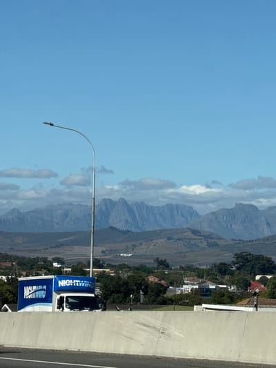 This image captures a wide, scenic view of a mountainous landscape, likely taken from an elevated vantage point such as an overpass. In the foreground, a light-colored concrete barrier or wall runs along the right side of the frame, with a segment of road visible beyond it. A tall, slender street lamp pole with a distinctive curved arm stands prominently on the left.
Partially visible behind the street lamp and the barrier is a white truck or van. Attached to the side of this vehicle, and also positioned lower in the frame, are two striking blue banners. Both banners prominently feature the word "NIGHTWING" in large, white capital letters. The lower banner also includes smaller, white text beneath "NIGHTWING," though it is less distinct.
The mid-ground reveals a sprawling valley characterized by fields and natural vegetation, appearing in shades of green and brown. Numerous houses and buildings, some with red roofs and others with grey, are scattered across this area, suggesting a residential or rural settlement.
Dominating the background is a majestic mountain range, stretching across the entire width of the scene. The mountains display rugged peaks and slopes in tones of blue and grey, indicating distance. Patches of white and grey clouds are interspersed among and above the mountain peaks, contrasting with the expansive, bright blue sky. The clear sky and strong illumination suggest it is daytime, likely late morning or early afternoon, with fine weather. The scene conveys a sense of peaceful natural beauty. No specific human activity or individuals are visible. The location context is Bellville, South Africa.