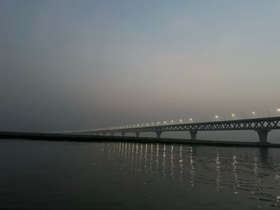 An elevated, multi-span truss bridge extends horizontally across a wide body of water, likely a river, in Naodoba, Bangladesh. The bridge features numerous evenly spaced streetlights illuminating its deck, which cast elongated, bright reflections onto the dark, rippled water below. A narrow strip of land or bank is visible between the water and the bridge structure on the left side of the frame. The sky above is a muted, hazy grey-blue, indicating either early morning, late evening, or overcast conditions, with the artificial lights providing the primary illumination. No individuals or active subjects are discernible in the scene. The bridge's structural elements, including its support pillars and diagonal cross-braces, are clearly visible against the subdued background.