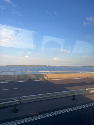 This media file shows a view from a bridge, looking down at a body of water and a road. The sky is a clear blue with some scattered clouds, suggesting it is daytime, likely in the morning or late afternoon due to the low angle of the sun casting long shadows. The water appears calm, reflecting the sky and some light, possibly indicating a mild weather day.
The bridge has a concrete barrier separating the roadway from the water, and a metal railing. The road itself is asphalt with white lane markings. There are lampposts or similar structures visible along the side of the road. The scene has a slightly reflective quality, possibly due to a window or a glass enclosure the photographer is situated behind, as evidenced by the faint reflections of clouds and rectangular shapes on the left side of the image.
The setting appears to be a developed area, given the presence of a bridge and a road, but the surrounding landscape is not clearly visible. The overall mood is serene and quiet, with no people or vehicles immediately apparent, focusing the attention on the infrastructure and the natural elements of the sky and water.