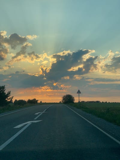 The image depicts a rural road at sunset in Niva, Russia. The sky is filled with dramatic clouds, illuminated by the setting sun, casting rays of light across the scene. The road curves to the right, with a white line marking the lane. To the left of the road, a field stretches into the distance, bordered by a line of trees. A triangular road sign is visible, indicating a sharp curve ahead. The overall atmosphere is serene and peaceful, with the warm hues of the sunset creating a sense of tranquility. The time of day is evident from the golden light and the long shadows. The weather appears clear, with the exception of the scattered clouds. No people are visible in the image, nor is there any visible text other than the road markings.