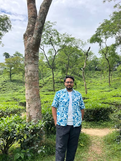 A young man stands on a dirt path in a tea garden in Sylhet, Bangladesh. He is wearing a blue and white patterned short-sleeved shirt, a white t-shirt, and black pants. He has glasses and is looking at the camera with a slight smile. The tea garden is lush and green, with rolling hills covered in tea bushes. Several trees are scattered throughout the garden, including a large tree in the foreground to the left of the man. The sky is overcast, with some clouds visible.