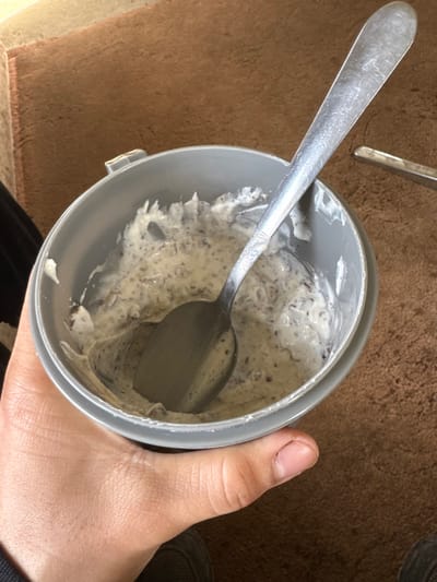 An overhead view depicts a light-skinned hand holding a round, light grey plastic container. Inside the container is a partially consumed white or off-white creamy substance heavily speckled with dark brown fragments. A silver-colored metal spoon, exhibiting surface scratches and a dull finish, is inserted into the substance, angled upwards and to the right. The hand holds the container from below, with visible knuckles and a short, natural thumbnail. A dark fabric item, likely clothing, is partially visible on the left side of the frame. The background consists of a textured, light brown surface, consistent with a carpet or rug. A portion of another metallic object is visible on the brown surface to the far right. The scene is located in Invergowrie, United Kingdom.