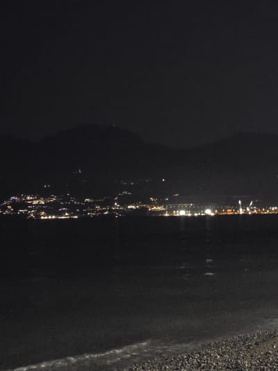 The image is a nighttime, wide-angle shot of the coastline of Salerno, Italy, as viewed from across the body of water. The foreground is dominated by a dark, pebbled beach. Beyond the beach, the dark water of the sea reflects the distant city lights. The middle ground features the vibrant, illuminated cityscape of Salerno, stretching along the coast and climbing the hillsides. Numerous lights, varying in color and intensity, indicate buildings, streets, and possibly a harbor area. In the background, dark, silhouetted mountains form the backdrop to the city. The sky is completely dark with no visible stars or moon. The overall impression is of a bustling city at night, seen from a peaceful, dark vantage point across the water.