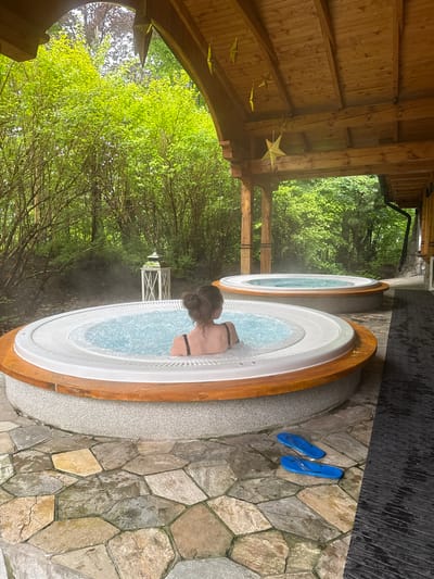 This vertical shot captures an serene outdoor spa setting in Zakopane, Poland, featuring two hot tubs nestled amidst lush greenery under a rustic wooden pergola. In the foreground, a person, viewed from behind, is relaxing in the bubbling hot water of one of the circular tubs, their dark hair tied up, wearing a black bikini top. The tub has a white textured interior and a warm wooden rim.
Beside the tub, on irregularly shaped stone paving, a pair of bright blue flip-flops are neatly placed, suggesting recent use. Another identical hot tub is visible just behind the first, also filled with water, hinting at more available relaxation.
The wooden pergola overhead features exposed beams and planks, decorated with several golden, star-shaped lanterns hanging down. Beyond the immediate spa area, dense foliage of various green trees dominates the background, creating a natural and secluded ambiance. A small white, possibly decorative, gazebo-like structure can be faintly seen through the trees in the distance. The scene is illuminated by natural daylight, suggesting a peaceful daytime experience. The steam rising from the hot tubs adds to the tranquil atmosphere. There is no visible text in the image.