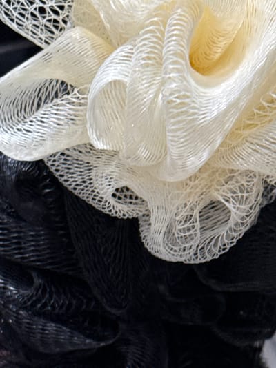 The media file features a close-up, abstract view of two loofahs, one white and one black, layered on top of each other. The white loofah, positioned on the left and center of the frame, has a soft, creamy texture with visible netting and swirling folds. The black loofah is visible on the right and bottom of the frame, appearing as a dark, textured mass with a similar netting structure. The lighting creates a soft focus, highlighting the textures of the loofahs. There is no discernible activity, scene, setting beyond the objects themselves, or time of day indicated. No text is visible. The location context of Likino-Dulyovo, Russia, does not provide any visual cues within the image.