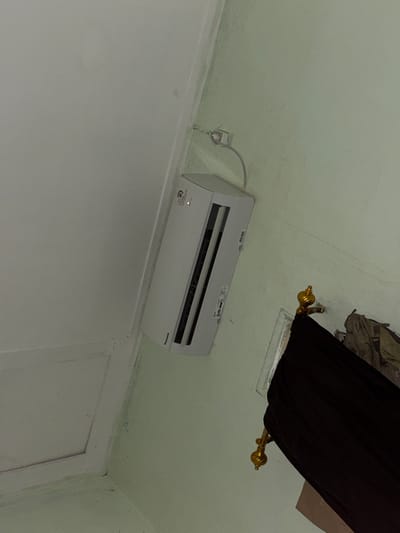 In a room, an indoor air conditioning unit is mounted high on a pale green wall. A white wire runs from the unit to a wall outlet. To the right of the air conditioner, a decorative gold finial is attached to a dark object, possibly part of a piece of furniture or a canopy. The wall appears to have some discoloration or mold in the upper right corner. The ceiling is white and shows a raised rectangular section. The overall scene suggests an indoor residential setting. The location is identified as Tupah, Indonesia.
