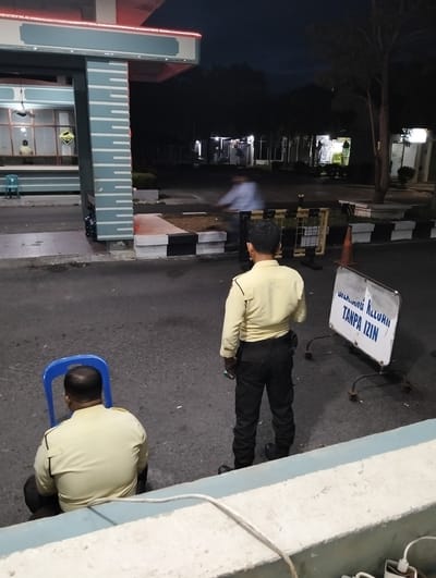 In the late evening or night, two security guards are present at an outdoor checkpoint or entrance in Tambontunong, Indonesia. The scene is dimly lit, with artificial lights illuminating the area.
One guard, wearing a light yellow uniform and dark pants, is standing and facing away from the camera, looking towards a road barrier. A white sign with blue text is visible to his right, which reads "DILARANG KELUAR TANPA IZIN" (No Leaving Without Permission). Another security guard, also in a similar uniform, is seated on a bright blue plastic chair, his back to the camera.
The setting appears to be a road leading to a building or complex. In the background, there are trees and several structures, some of which are illuminated. A blurred figure on a bicycle or motorcycle is visible in the middle ground, suggesting some ongoing traffic. The overall atmosphere is calm and functional, typical of a security post at night. The weather appears to be clear, with no visible rain.