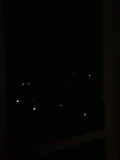 The image depicts a night scene in Krykhivtsi, Ukraine. It is taken from inside a building looking out through a window. The outside is dark, with scattered points of light that could be city lights or stars. The window frame is visible on the right side of the frame. The lighting conditions are extremely low, and details are obscured.
