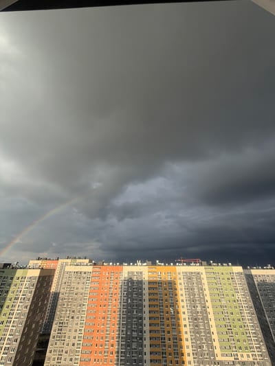 The image depicts a landscape in Анкудиновка, Russia. A large, multi-story building dominates the right side of the frame. The building has a varied color scheme with alternating horizontal bands of grey, orange, and green. The facade of the building is punctuated by rows of windows. To the left of the building is a cloudy, grey sky with a faint rainbow arching across the lower portion.