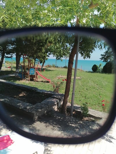 This media file shows a view through eyeglasses onto a playground and the sea. The foreground is dominated by the dark rim of the eyeglasses, with shadows of leaves cast on a paved area and a white object that appears to be a lounge chair with a red towel.

Through the lenses, a playground structure with a red slide, swings, and a climbing net is visible on a grassy area. In the background, a vast expanse of calm, turquoise sea stretches to the horizon under a clear, bright sky, suggesting a sunny day. The presence of trees with lush green foliage framing the scene indicates a warm, possibly summer, season. Red roses bloom near a stone planter in the middle ground, adding a touch of color to the grassy landscape. A thin metal pole stands near a tree, and a red marker pole is visible in the distance near the water's edge.

The scene evokes a sense of leisure and outdoor recreation, likely a holiday setting. The location is identified as Durankulak, Bulgaria. No people are visible in the image, but the playground implies children's activity. The overall mood is peaceful and bright. No visible text is present in the image.