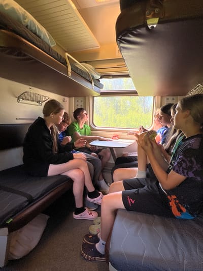 The image depicts a group of five young people inside a train compartment. The compartment has bunk beds on the upper level. The people are seated on the lower bench, facing a table positioned by the window. Sunlight streams through the window, illuminating the interior. One individual, on the far left, is wearing a black hoodie and shorts. Next to them, a person in a striped shirt is visible. Another person in a green t-shirt is looking out the window. To their right, two individuals are seated, with one holding some cards. The floor of the compartment is carpeted, and various footwear, including Crocs, are visible. The environment suggests a journey on a train, potentially in or near the city of Nizhniye Vyazovye, Russia, given the context provided.