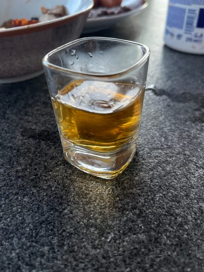 A close-up shot focuses on a single shot glass filled with amber-colored liquid, likely alcohol, placed on a dark, speckled granite surface. The glass is square-shaped with rounded edges and appears to have condensation on its exterior, indicating it is cool. The liquid inside is clear and reflects the light, creating a golden hue. In the background, out of focus, is a portion of a bowl containing food items and what appears to be a white container with blue text. The lighting suggests an outdoor setting, possibly a patio or table on a sunny day, with the sun casting subtle shadows. The scene is devoid of any human presence. This scene does not appear to be in Milná, Czechia.