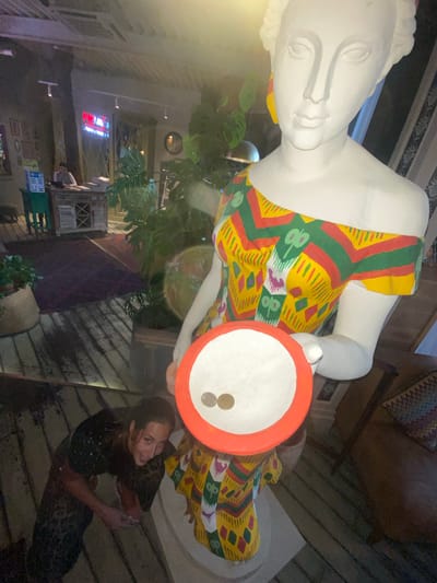 In Solnechnoye, Russia, a woman kneels on a wooden deck, looking up and smiling at a large, white statue of a woman. The statue wears a colorful, patterned dress and holds a white bowl with an orange rim, containing two coins. Plants are visible in the background, along with a reception desk.