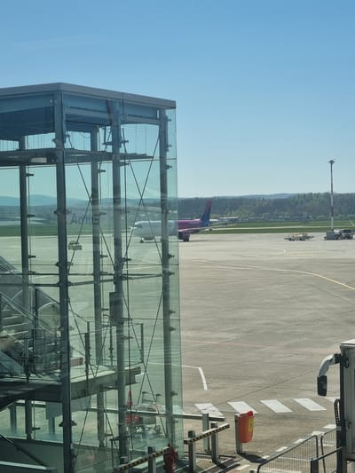 The image shows an airport scene viewed from a glass-walled structure, likely a jet bridge. An airplane with a distinctive purple tail is parked on the tarmac, which is marked with yellow and white lines. Various airport vehicles and equipment are visible in the background. The sky is clear and blue. Based on the location, this scene is likely at EuroAirport Basel-Mulhouse-Freiburg, which is partially located in Saint-Louis, France.
