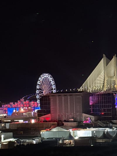 The image depicts an outdoor event at night, likely a festival or concert, held near the Vasco da Gama Bridge in Loures, Portugal. A large Ferris wheel, illuminated with white lights, is prominently visible on the left side of the frame. The Vasco da Gama Bridge itself, a cable-stayed bridge, is illuminated with white lights, highlighting its distinctive architecture. In the foreground, various temporary structures, including tents and stages, are set up. Some of these structures are also lit, casting a warm glow on the scene. The overall atmosphere appears festive and lively, with the bright lights contrasting against the dark night sky. There are a few faint green lights visible in the sky, possibly drones or distant aircraft.
