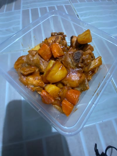 A close-up, top-down shot shows a clear plastic food container filled with a savory dish. The dish consists of pieces of chicken, some with bone, coated in a rich, glossy brown sauce. Interspersed with the chicken are chunks of bright orange carrots and golden-yellow potatoes, also coated in the sauce. The food appears to be freshly cooked and has a appetizing look. The container is placed on a tiled surface with a light blue and white pattern, suggesting an indoor setting. The lighting is dim, casting shadows on the tiled surface and on the food itself, creating a cozy and perhaps domestic atmosphere. The scene appears to be in San Jose del Monte, Philippines, implying a local Filipino dish, possibly adobo or a similar stew. There is no visible text in the image.