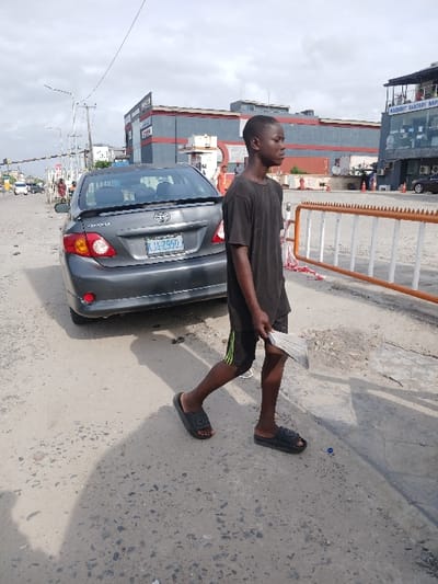 A young man wearing a black t-shirt and black shorts, walks on a paved road in front of a grey Toyota Corolla. The man is holding a piece of paper in his right hand. He is wearing black sandals. The car has a license plate that reads "KJA-2950." There is a building in the background with a sign that says "Midland Sanitary Mart." The road is rough and pockmarked. A blue bottle cap is on the ground to the right of the man. The scene is in Way, Nigeria, located at latitude 6.429, longitude 3.468.