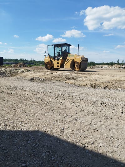 A yellow Caterpillar roller compactor, identified by the "CAT" logo on its side, is positioned on a construction site in Rozdil, Ukraine. The compactor is oriented diagonally from left to right, with its front drum facing towards the viewer's left. The machine has a glass-enclosed cabin. The ground is covered with gravel and dirt. In the background, there are piles of earth, some wooden pallets, and a line of trees. The sky above is blue with scattered white clouds. The overall scene depicts an active construction area under a clear sky.