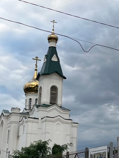 The image shows a white church with golden domes against a cloudy sky. The church has multiple levels and a green roof. Power lines are visible in the foreground. A fence and a sign with text are present at the bottom right of the image. The sign is in Russian and appears to be an advertisement or informational poster.