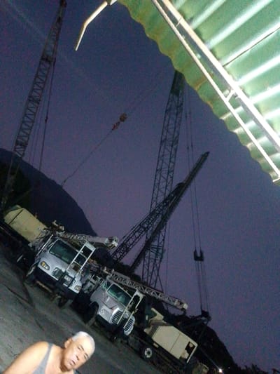 The image shows a man wearing a tank top in the foreground. Behind him are two crane trucks parked on a paved surface. In the background, two large cranes are visible against a dusk sky. A hillside is also visible in the background. The location is Parroquia Guatire, Venezuela.