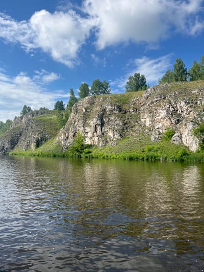 This image captures a scenic view of the Iset River in Kamensk-Uralsky, Russia. The river occupies the foreground, its surface rippling and reflecting the light. The background features a steep, rocky cliff face overgrown with lush green vegetation, including trees and grass. The top of the cliff is dotted with more trees. The sky above is a vibrant blue, with scattered white clouds. The scene conveys a sense of natural beauty and tranquility.