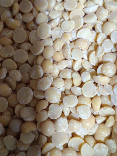The image is a close-up, overhead shot of a large pile of split peas. The peas are a light yellow-green color, with some appearing more yellow than others. They are irregularly shaped, as is typical for dried split legumes, and some show a rough texture. The lighting is somewhat dim, creating shadows that add depth and highlight the uneven surface of the pile. The background is completely filled with the peas, with no discernible environmental features or context. There are no people, actions, or interactions visible in the image. It's a static, textural study of dried split peas.
