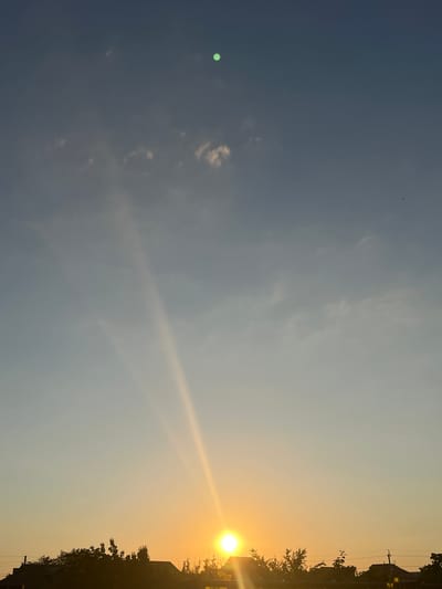 The image depicts a clear sky with a soft gradient of blue to yellow, indicating either sunrise or sunset. The sun, a bright orb of golden light, is positioned near the horizon in the lower right portion of the frame. Sunbeams fan out from the sun, creating dramatic rays that stretch upwards into the sky. Silhouetted against the bright sky are the rooftops of several houses and the dark shapes of trees, suggesting a residential area. A few small, wispy clouds are scattered across the upper sky. The overall impression is one of tranquility and the natural beauty of the sky. The context provided indicates the location is Slobozia, Moldova.
