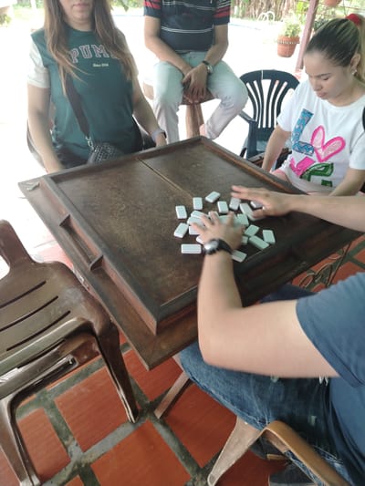 A group of four people are sitting around a wooden domino table outside in Los Leoncitos, Venezuela. One woman is wearing a Puma t-shirt with a shoulder bag. Another woman is wearing a white shirt with heart graphics. One man is wearing a striped shirt, and another is in a blue shirt, gathering dominoes on the table. The table sits on a red tile floor.