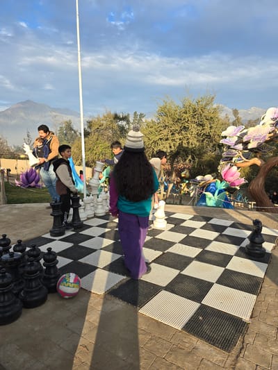 The image shows a group of people on a giant chessboard in an outdoor setting. A woman with long dark hair and a striped hat stands on the board, wearing a teal shirt and purple pants. Several other people, including children, are situated near the chessboard, and some are standing on large chess pieces. The chessboard itself is made of black and white squares. In the background, there are trees, a mountain range, and a blue sky with clouds. Artificial flowers and other decorations are also visible. A multi-colored ball is placed next to a group of black chess pieces. Based on the presence of mountains and specific landmarks, the image likely originates from the city of Puente Alto, Chile.