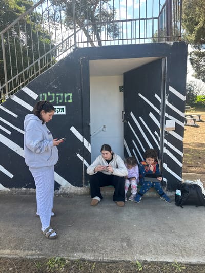 Four individuals are positioned outside a concrete structure in Beit Yitshak Sha'ar Hefer, Israel. The structure is painted black with diagonal white stripes and features an open doorway revealing a white interior and downward-leading stairs. A metal railing is visible above the structure. A green sign displaying "מקלטים" (shelters) in Hebrew is affixed to the black wall.
From left to right:
1. A female, appearing to be a young adult, stands on a concrete surface. She wears a light grey hooded sweatshirt, light-colored patterned pants, and brown sandals with white socks. She holds and interacts with a smartphone.
2. A second female, also a young adult, is seated on the concrete threshold of the open doorway. She wears a cream-colored long-sleeved top, dark loose-fitting pants, and light brown slip-on shoes. She holds and interacts with a smartphone.
3. A young girl, a child, is seated to the right of the second female. She wears a light purple long-sleeved top, light-colored patterned pants, and light blue sneakers. She holds and interacts with a small tablet device.
4. A young boy, a child, is seated to the right of the young girl. He wears a multi-colored long-sleeved top, dark patterned pants, and blue sneakers. He holds and interacts with a larger tablet device, with one hand near his mouth.
A black backpack and a white bag rest on the concrete surface to the right of the seated boy. The surrounding environment includes patchy grass and dirt, with several trees in the background under a partially cloudy blue sky. Wooden stump-shaped seating is visible in the distant background to the right. All four individuals are focused on their respective electronic devices.